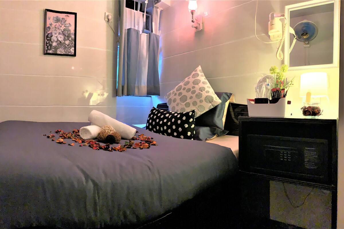 A neatly made bed with a grey bedspread, two pillows, and a rolled-up towel. A small table with a vase of flowers and a lamp is beside the bed.