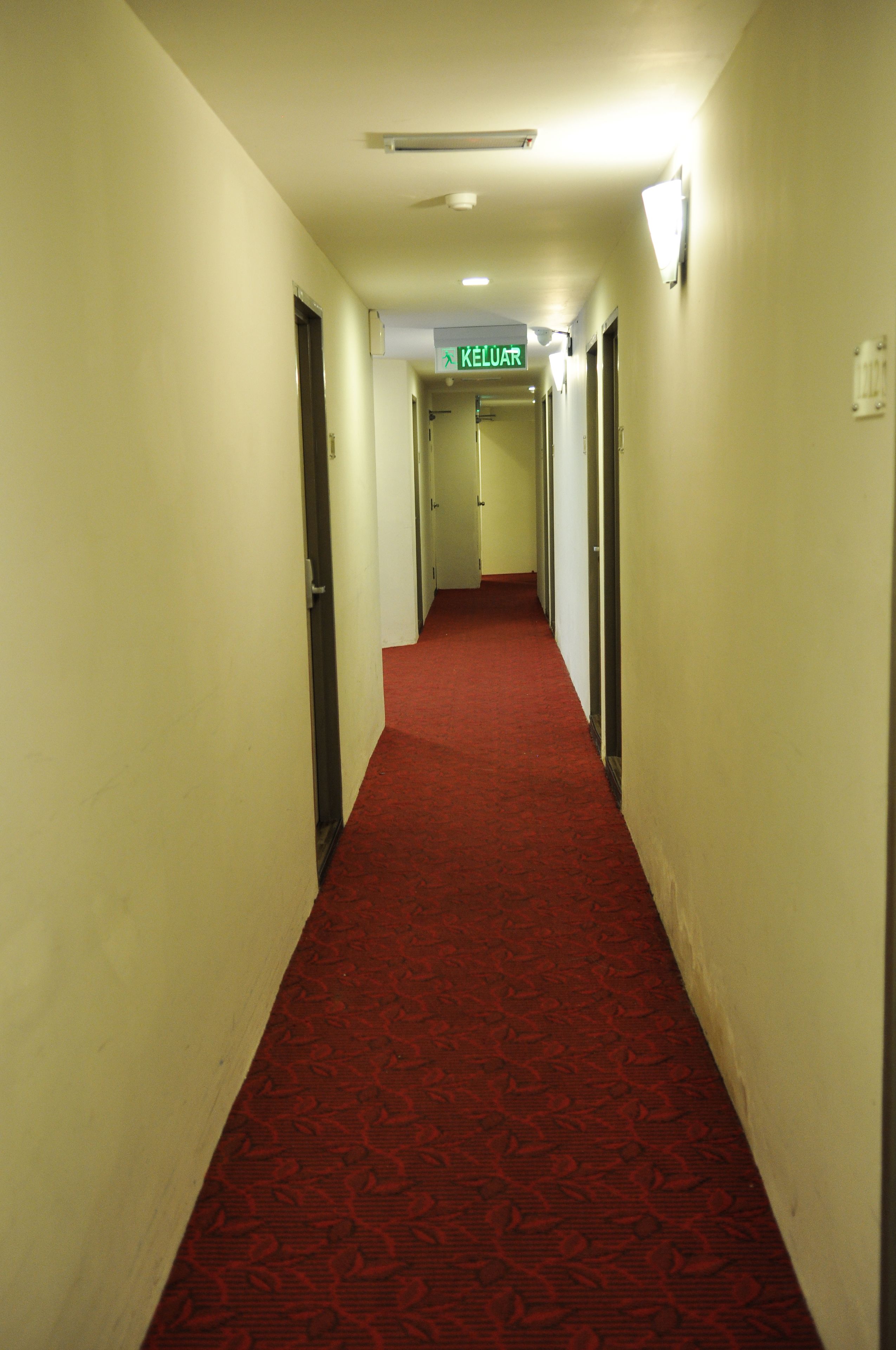 A hotel hallway with a red carpet, doors on both sides, and a green exit sign.