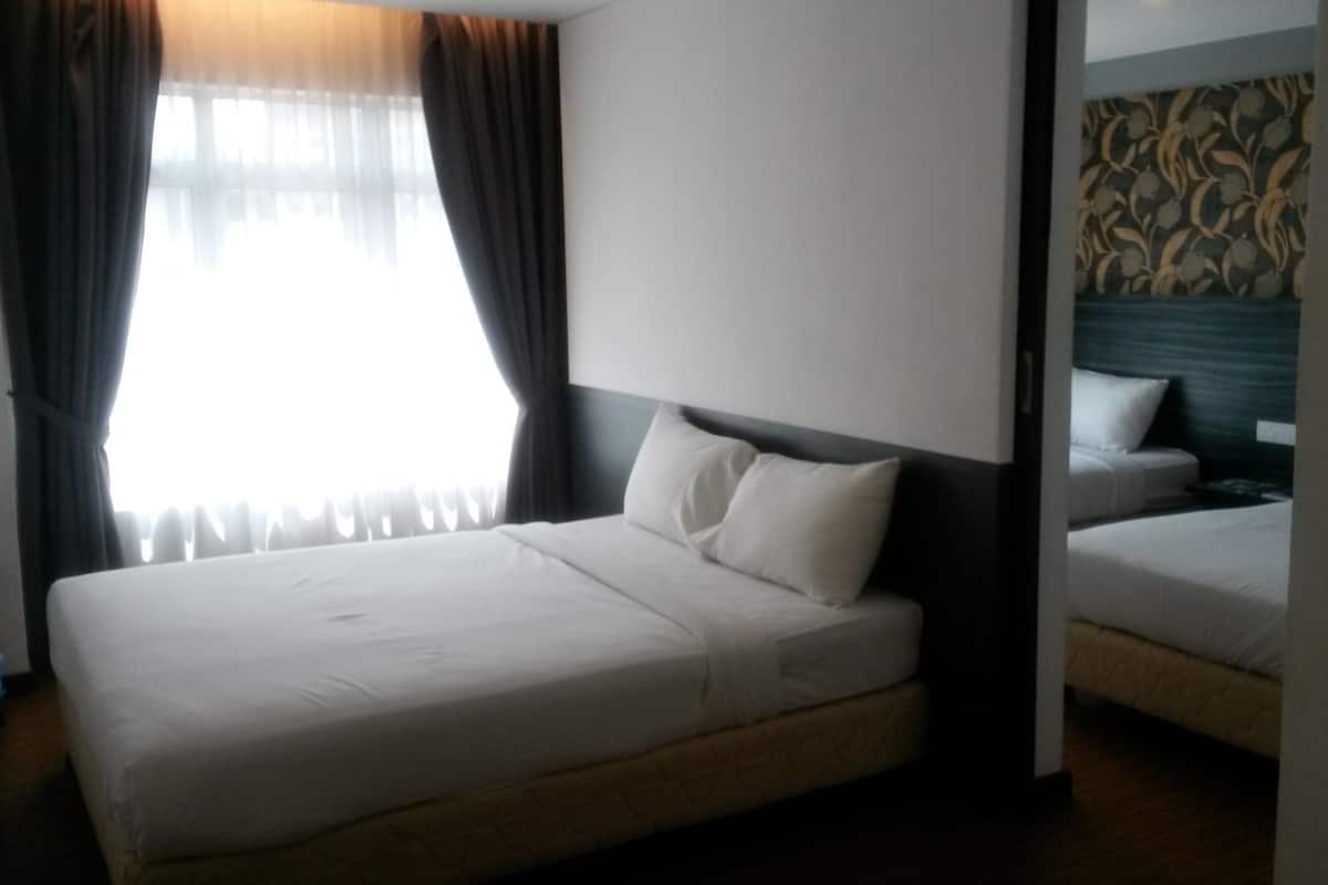 Quarto de hotel com uma cama de solteiro, uma segunda cama ao fundo, uma janela grande com cortinas e uma parede com padrão.