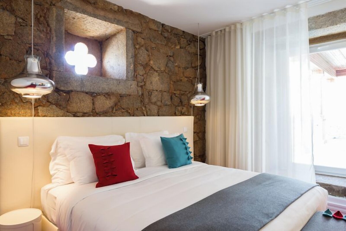 Una cama con ropa de cama blanca y una manta gris, pared de piedra con ventana y dos lámparas colgantes.