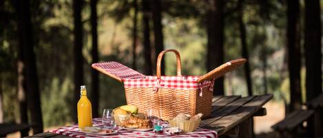 A picnic setup with a basket, fruits, bread, and a bottle on a wooden table in a forest.