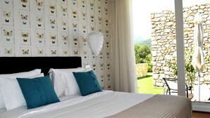 Habitación de hotel con una cama grande, ropa de cama blanca, almohadas color esmeralda, un adorno de pared con plumas blancas y vistas a un muro de piedra y follaje exterior.