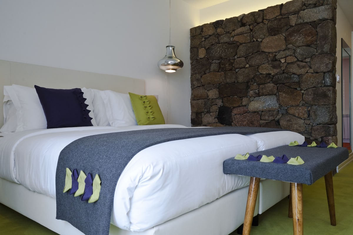 A bedroom with a stone wall, a bed with white bedding, a grey blanket, and a bedside table with a pillow.