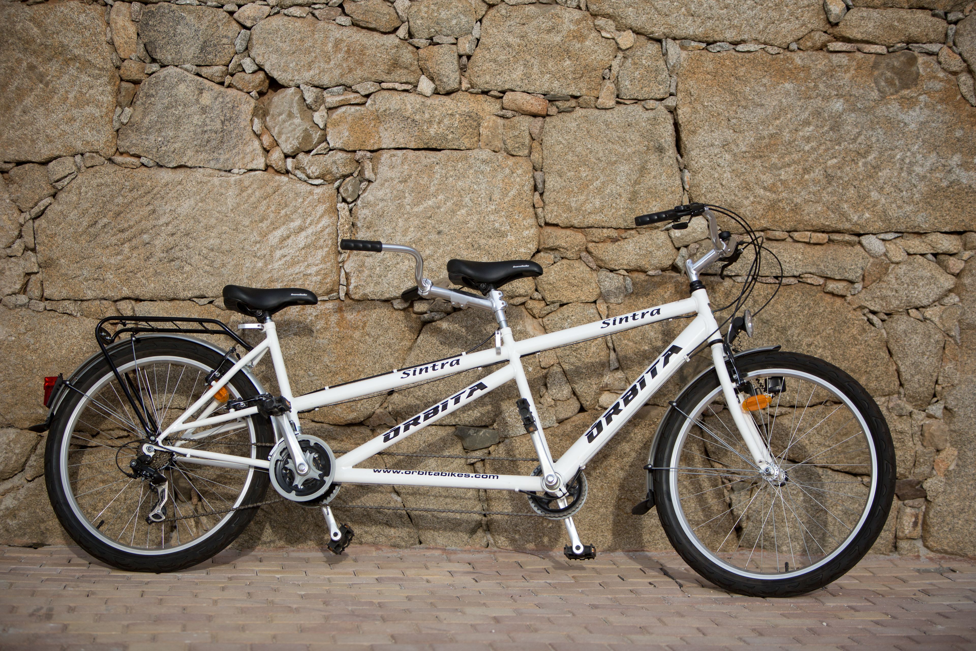 Una bicicleta blanca con los nombres de marca 'Sintra' y 'Orbitia' a la vista, apoyada contra una pared de piedra.