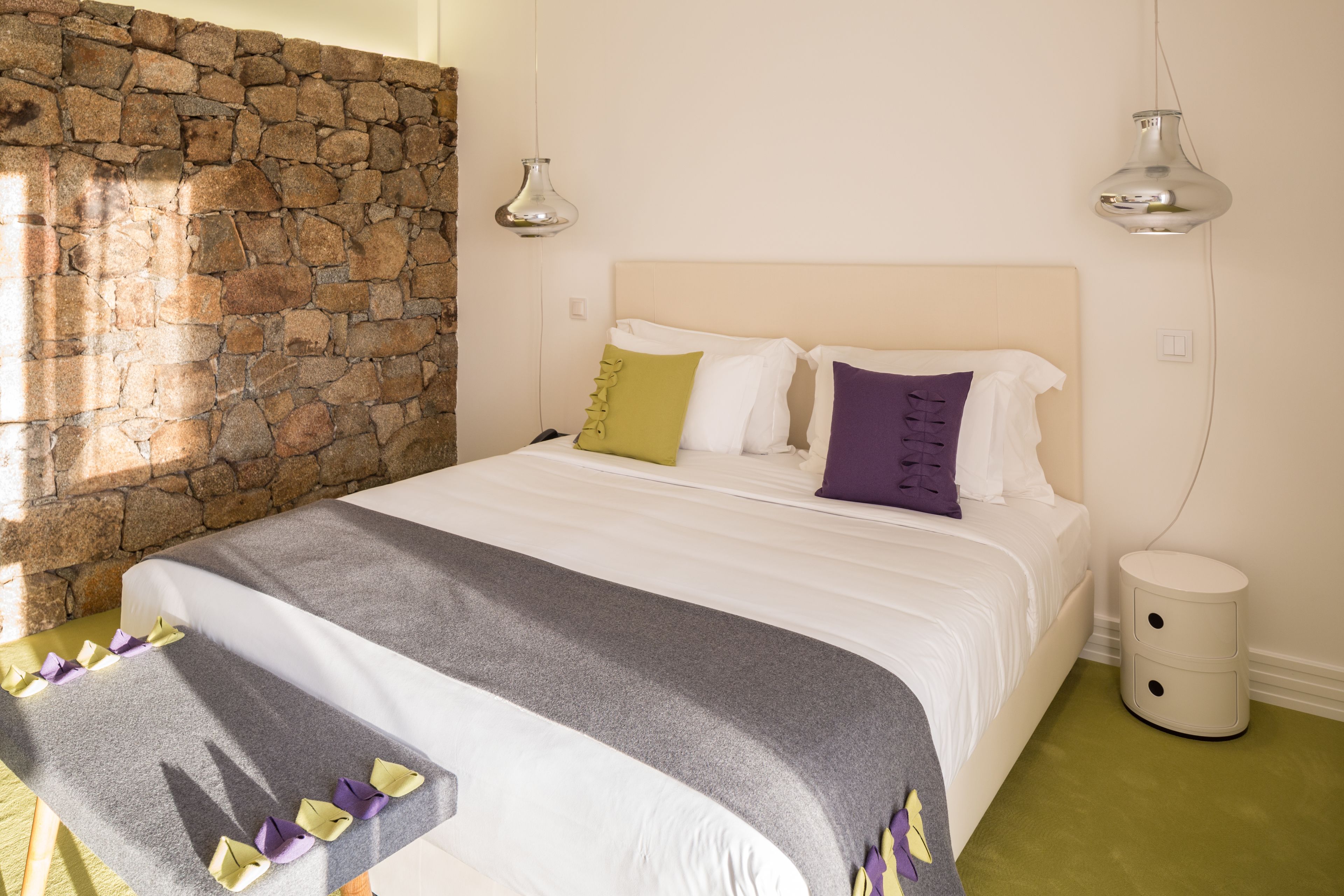 Un dormitorio con pared de piedra, una cama con edredón gris y blanco, y dos lámparas de noche.
