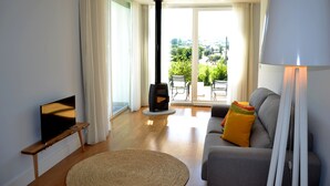 Una sala de estar con una televisión sobre un mueble de madera, un sofá con cojines, una lámpara, una alfombra y vistas a un área exterior con una silla y una mesa.