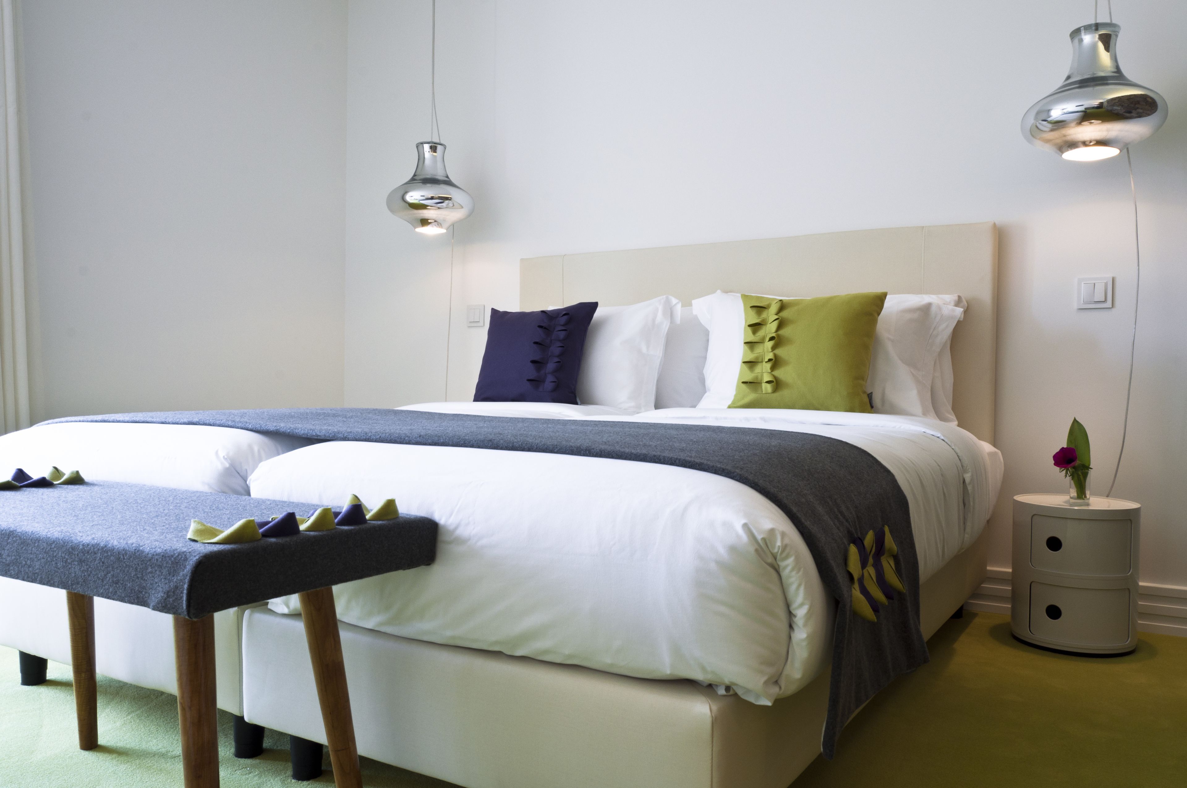 A neatly made bed with a grey and white duvet, a bedside table with a vase of flowers, and two pendant lights above the bed.