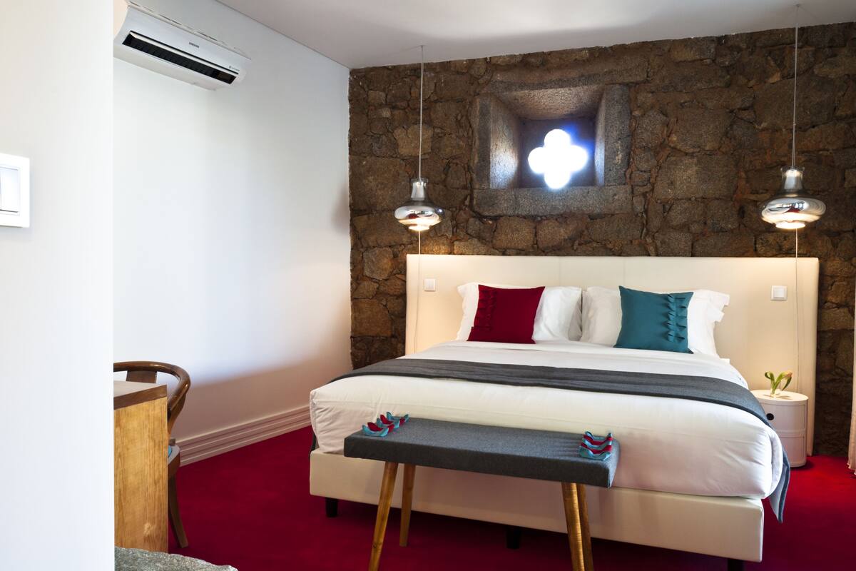 A bedroom with a stone wall, a bed with white bedding and red and teal pillows, a bench at the foot of the bed, and a small bedside table with a plant.
