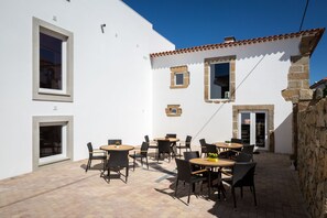 An outdoor patio with multiple tables and chairs, a stone wall, and a white building with windows and doors.