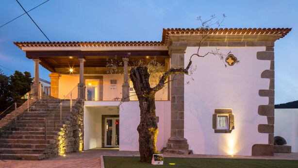 Un edificio de dos pisos con una escalera de piedra que lleva a la entrada, un gran árbol en frente y un tejado de tejas.