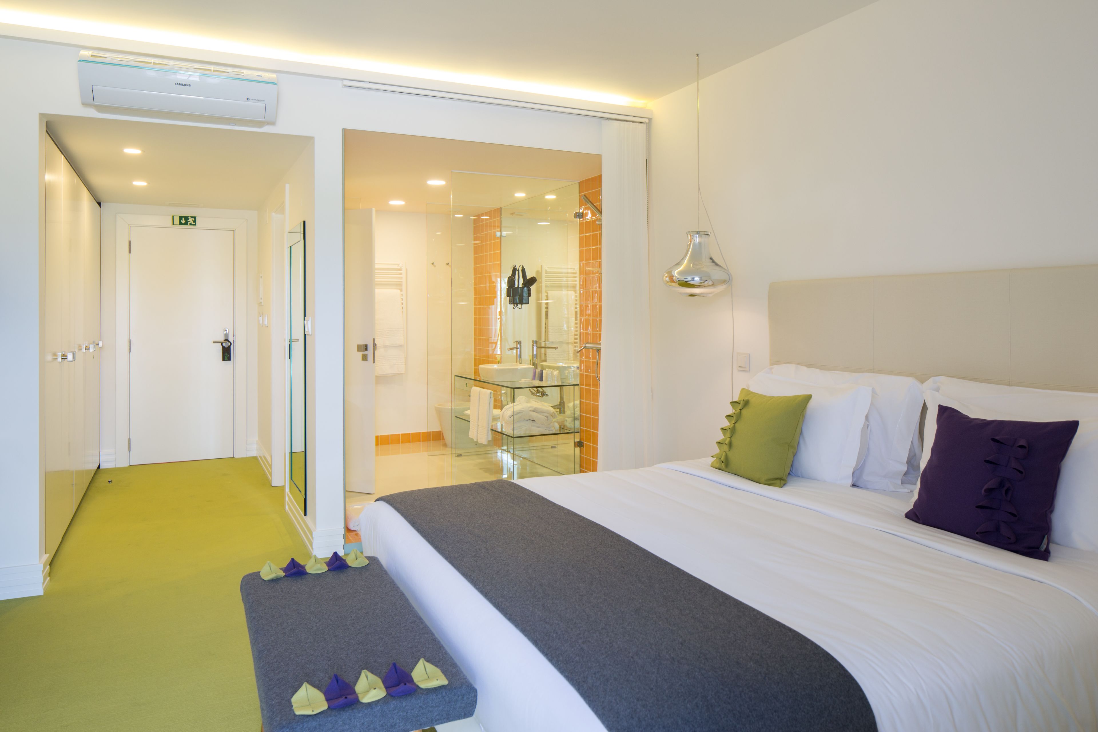 A hotel room with a bed, a glass shower area, and a hallway leading to an exit.