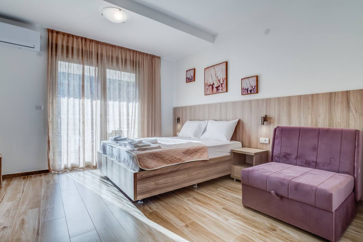 Quarto moderno com cama de madeira, sofá roxo e uma grande janela com cortinas leves.