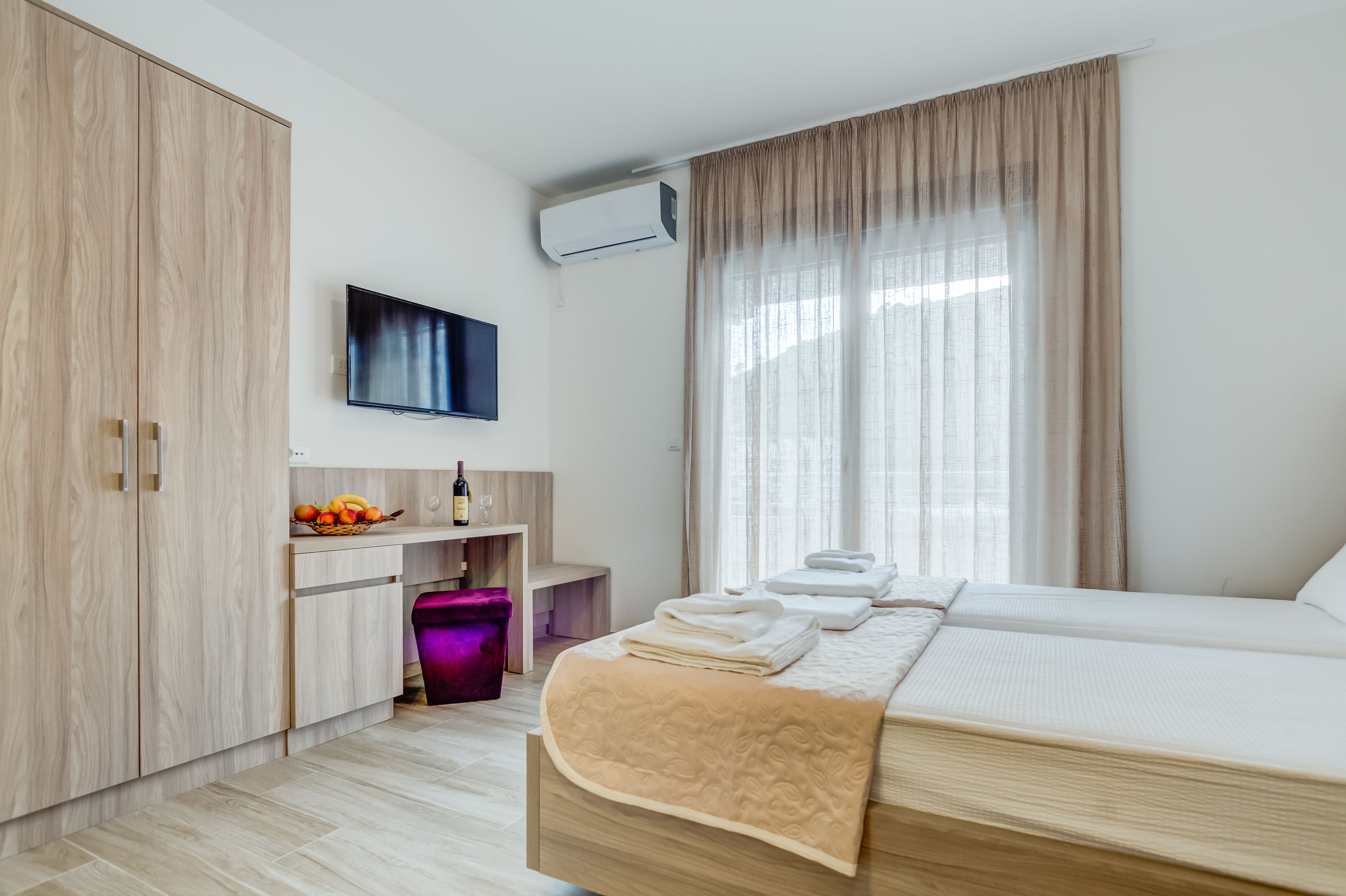 A modern hotel room with a large bed, a flat-screen TV, a minibar, and a large window with curtains.