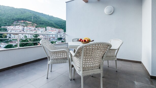 A balcony with wicker chairs, a table with fruit, and a view of a town and hills. - Adriatik Budva apartments (Budva)
