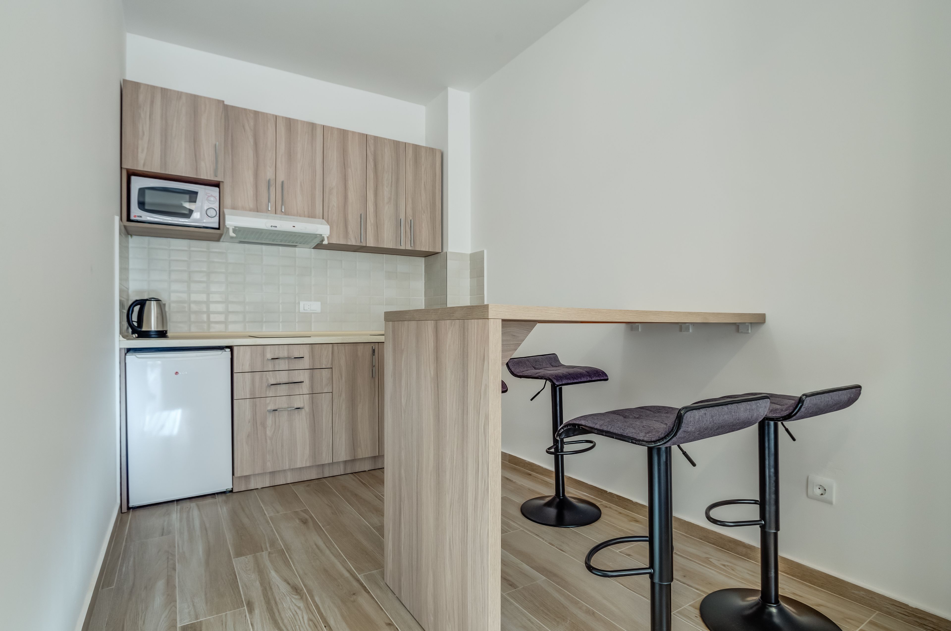A compact kitchen with a microwave, kettle, and a small refrigerator. There are two bar stools with cushioned seats.