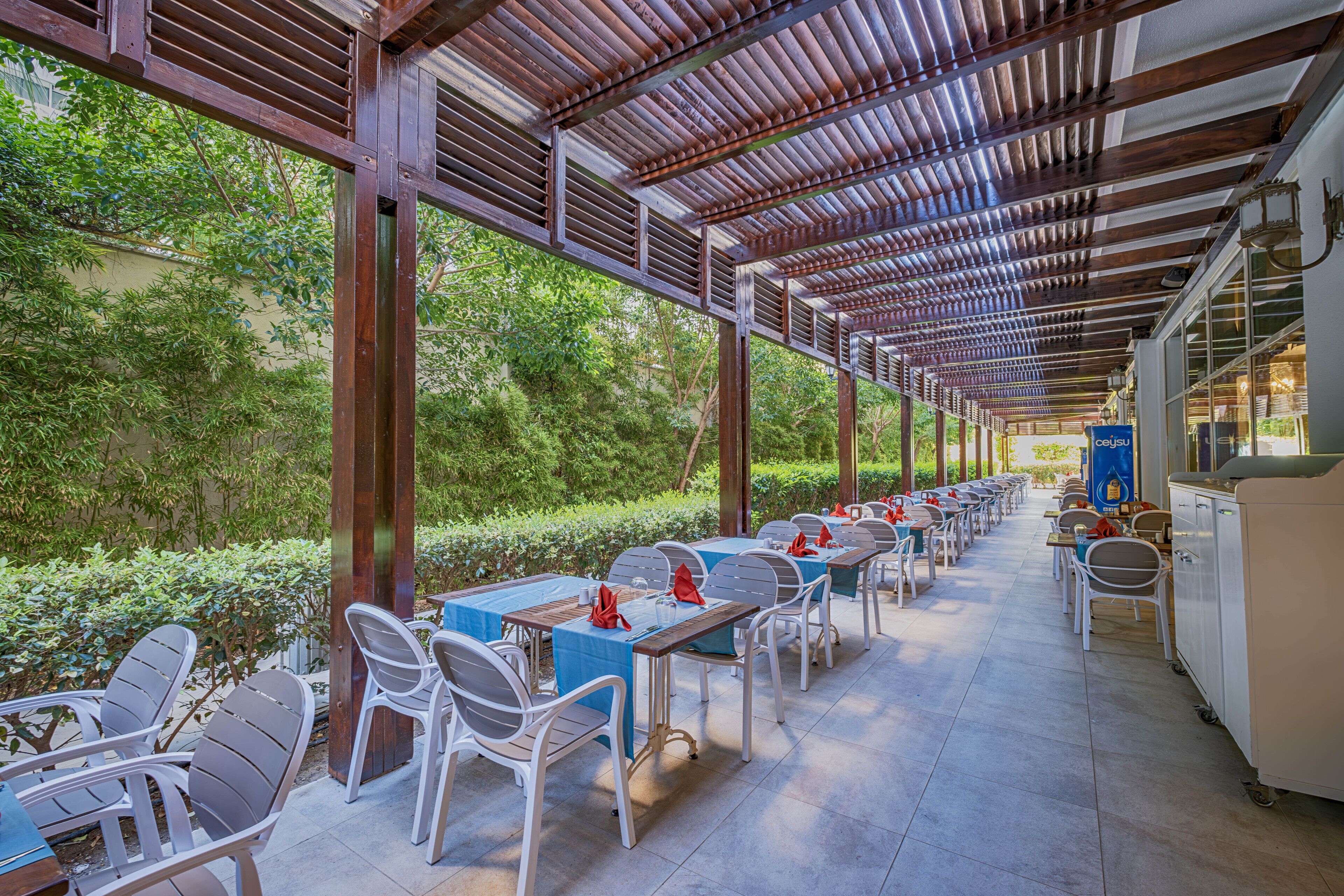 Un área de comedor al aire libre con mesas y sillas, techo cubierto y vistas a la vegetación.