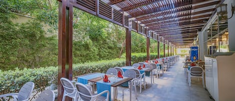 Un área de comedor al aire libre con mesas y sillas, techo cubierto y vistas a la vegetación.