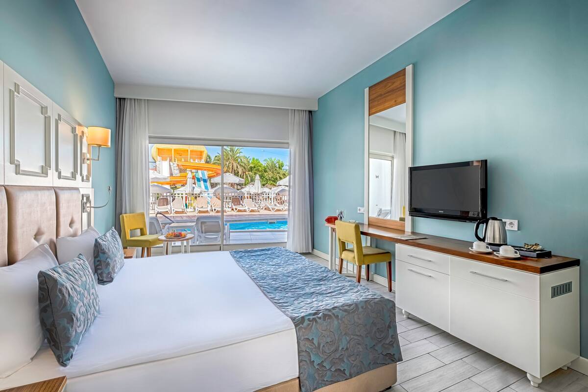 A hotel room with a bed, a desk, a TV, and a view of a water park.