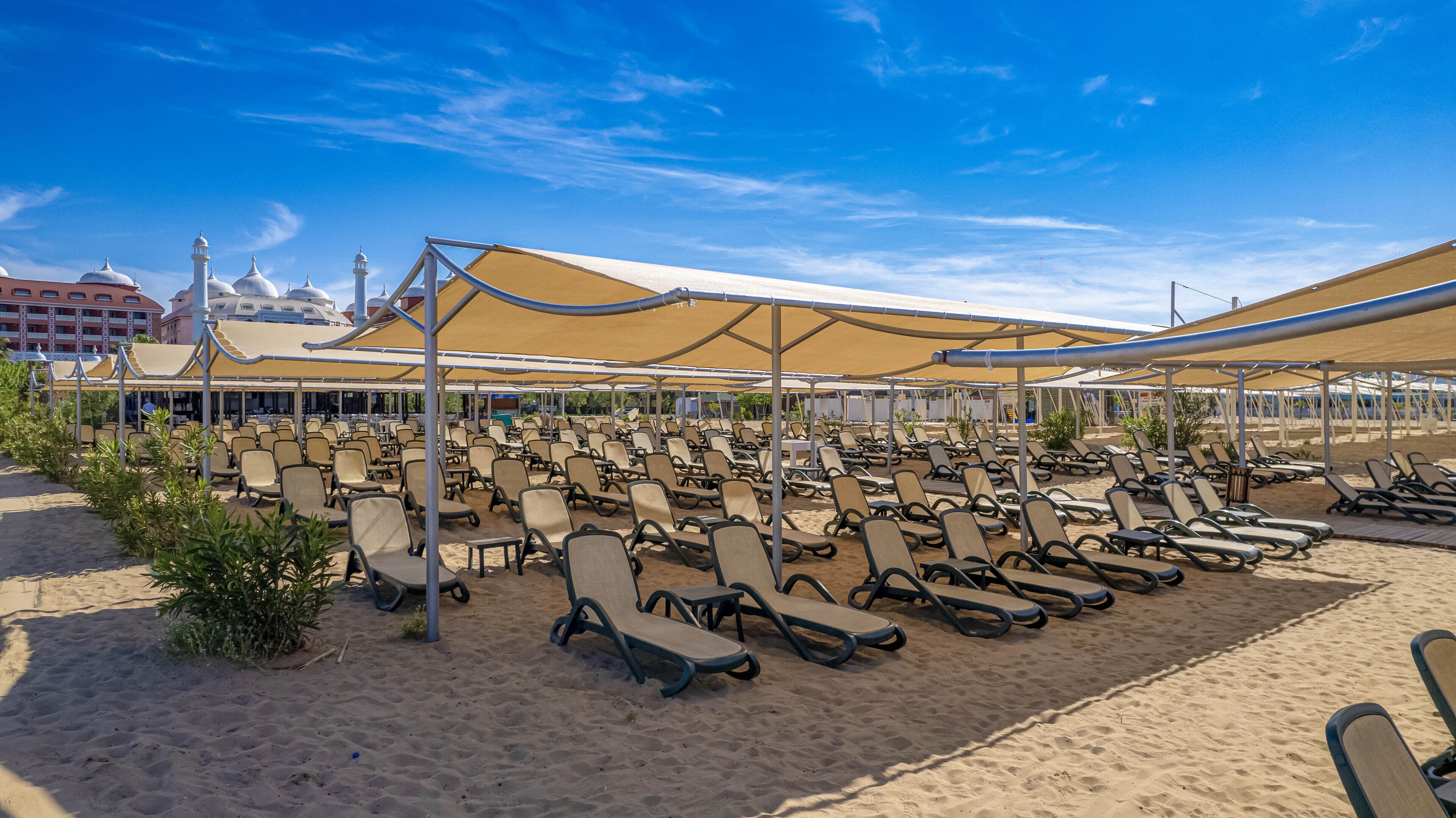 Una zona sulla spiaggia con file di lettini sotto grandi ombrelloni, una spiaggia sabbiosa e un edificio con architettura a cupola sullo sfondo.