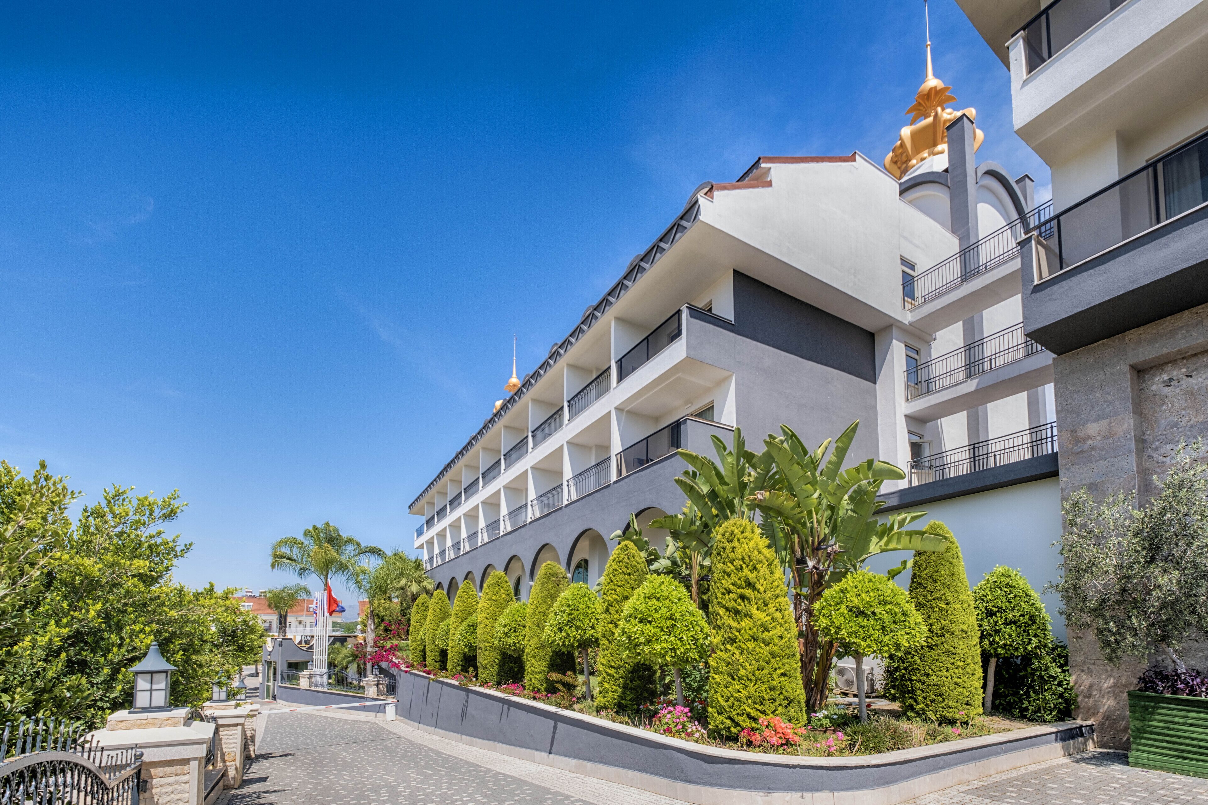 Un hôtel à plusieurs étages au design moderne, doté de balcons, d’une cour intérieure agrémentée d’arbres aux formes soignées et surplombant un ciel d’un bleu éclatant.