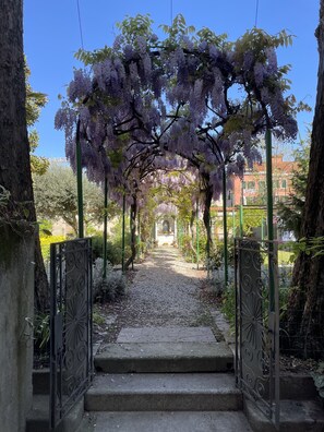 A pathway leading through a garden with purple flowering trees and a wrought iron gate. - Casa Caburlotto (Venice)