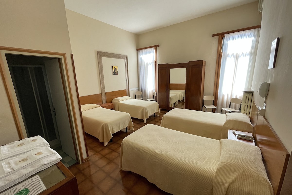 A hotel room with two single beds, a wooden wardrobe, a mirror, a small table with a chair, and a window with curtains.