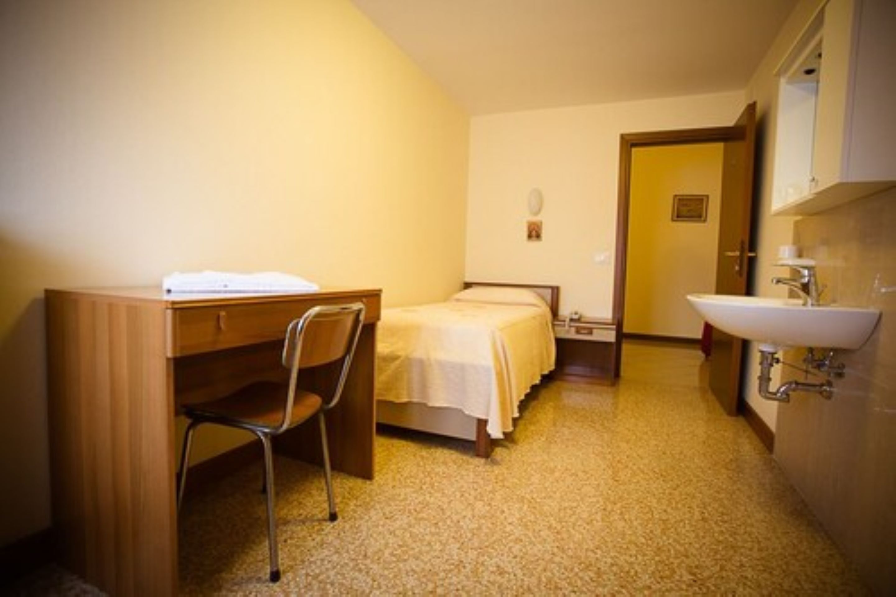 A small, compact room with a wooden desk, a single bed, a chair, and a bathroom with a sink and mirror.