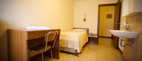 A small, compact room with a wooden desk, a single bed, a chair, and a bathroom with a sink and mirror.