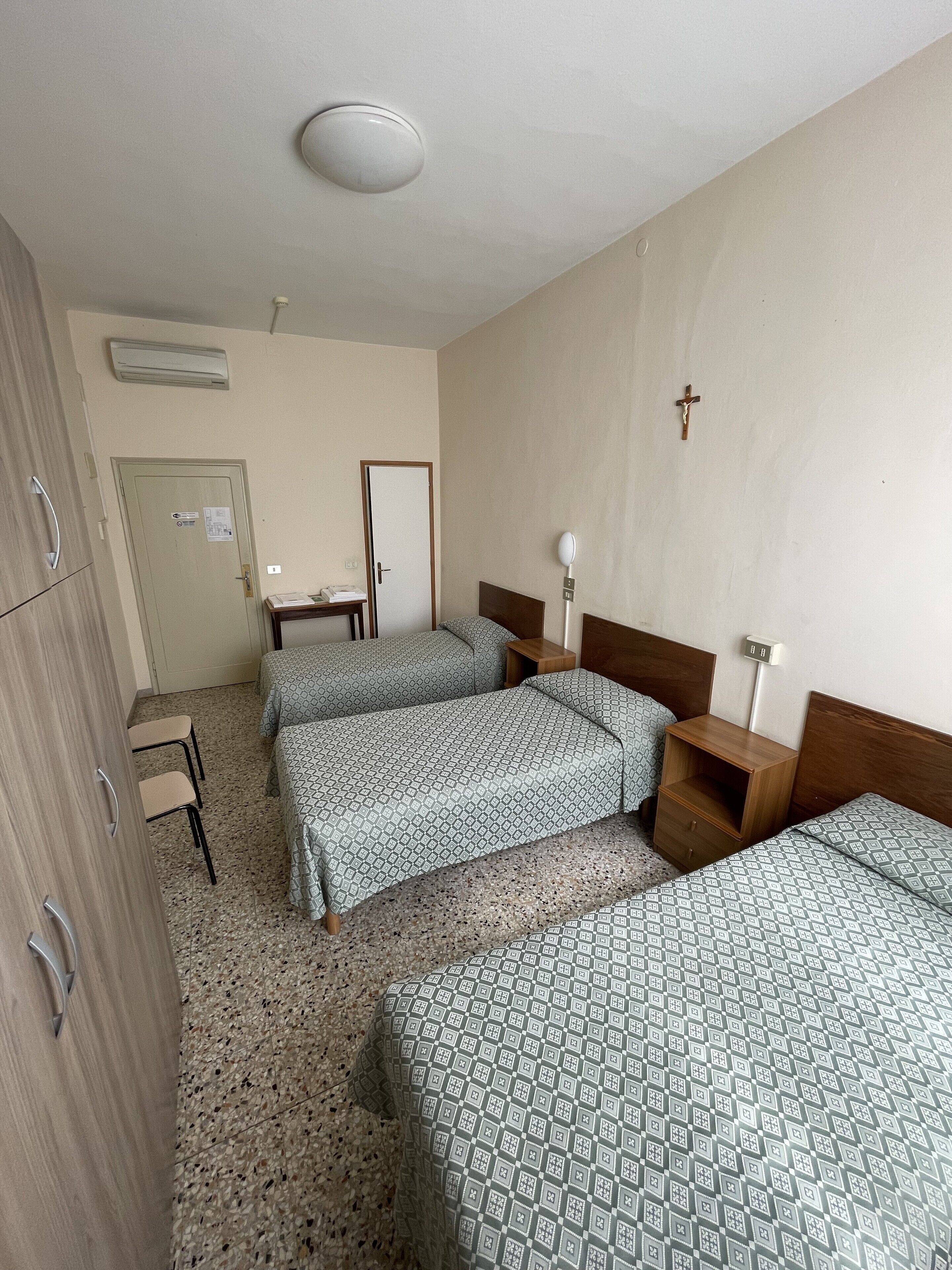 A hotel room with three beds, a wooden wardrobe, a desk with a chair, and a wall-mounted air conditioner.
