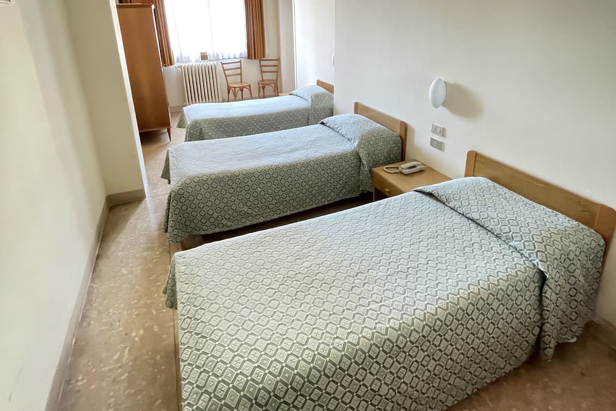 A hotel room with three single beds, a wooden headboard, a bedside table with a lamp, and a view of a dining area with a table and chairs.