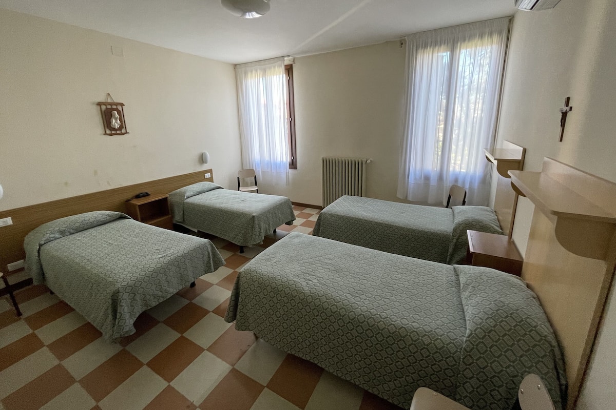 A hotel room with four single beds, a radiator, a window with curtains, a wall-mounted lamp, and a small table.