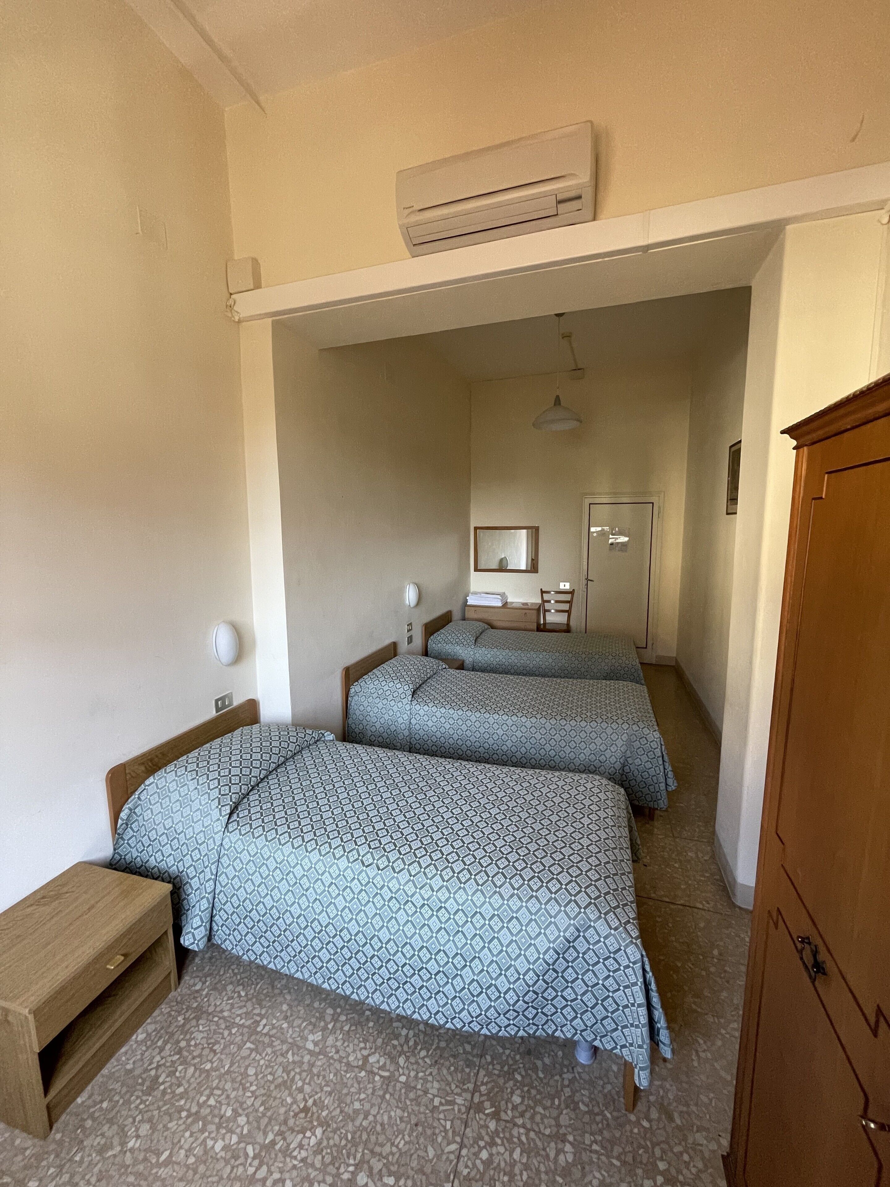 A room with two beds, a wooden cabinet, a desk, a mirror, and an air conditioning unit.