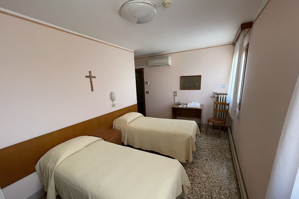 A hotel room with two single beds, a desk with a chair, a cross on the wall, and an air conditioning unit.