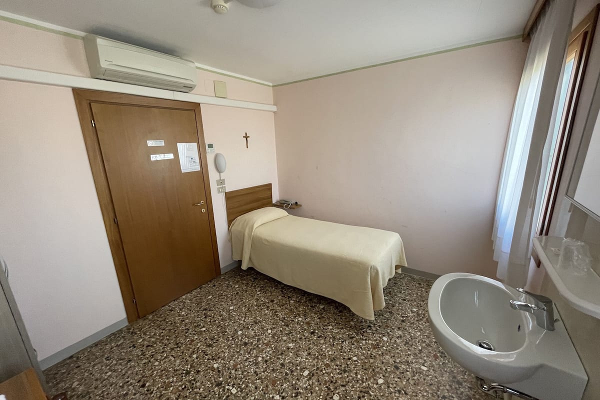 A small, clean room with a bed, a wooden door, a wall-mounted air conditioner, a cross decoration, a sink, and a mirror.