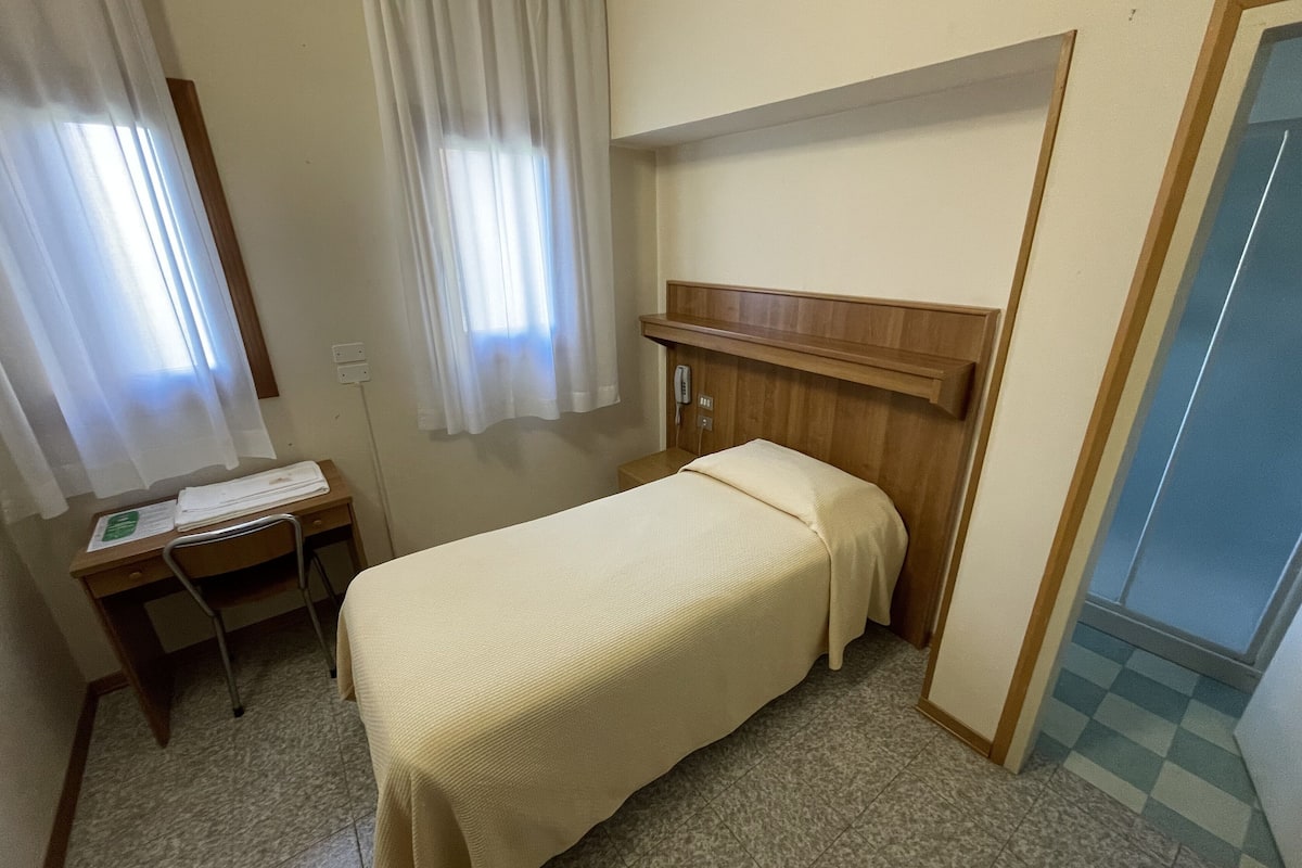 A small, single-bed room with a wooden headboard, a desk with a chair, a telephone, and a window with curtains.