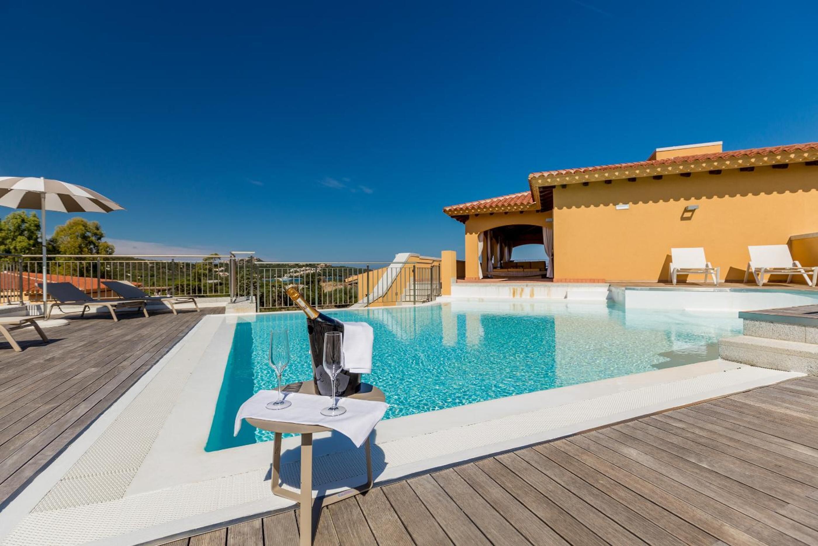 Una piscina en la azotea con sillones de descanso, una mesa con una botella y vasos, y un edificio con tejado de baldosas.