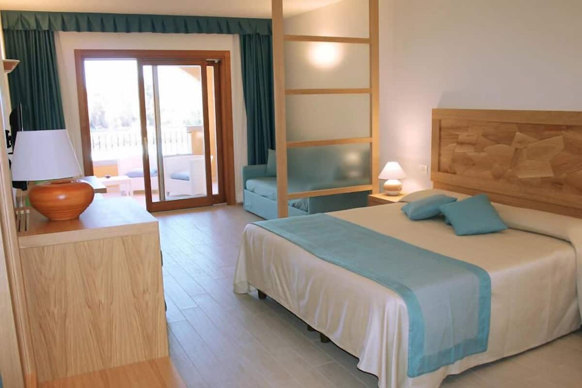 A hotel room with a bed, a desk with a lamp, a chair, and a view of the outside.