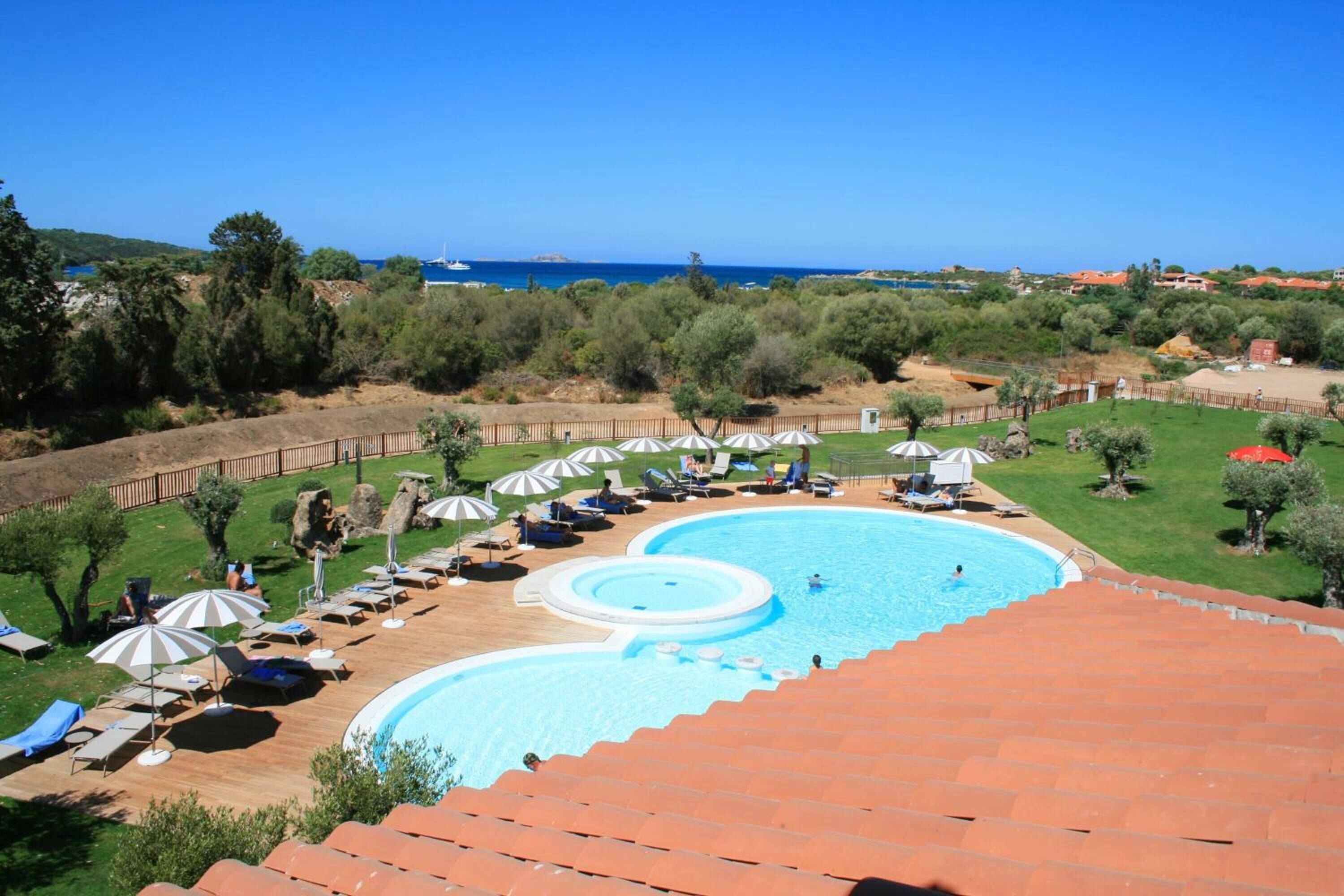 Ein Schwimmbecken mit Liegestühlen und Sonnenschirmen, umgeben von Grünflächen und Blick auf das Meer.
