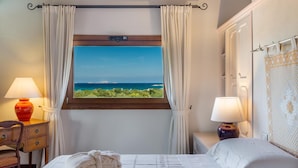 A bedroom with a bed, two bedside tables, a window with curtains, and a view of the sea. - Hotel Marana (Golfo Aranci)