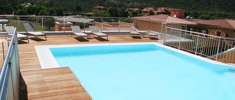 A pool area with lounge chairs and a wooden deck, surrounded by buildings and hills in the background.