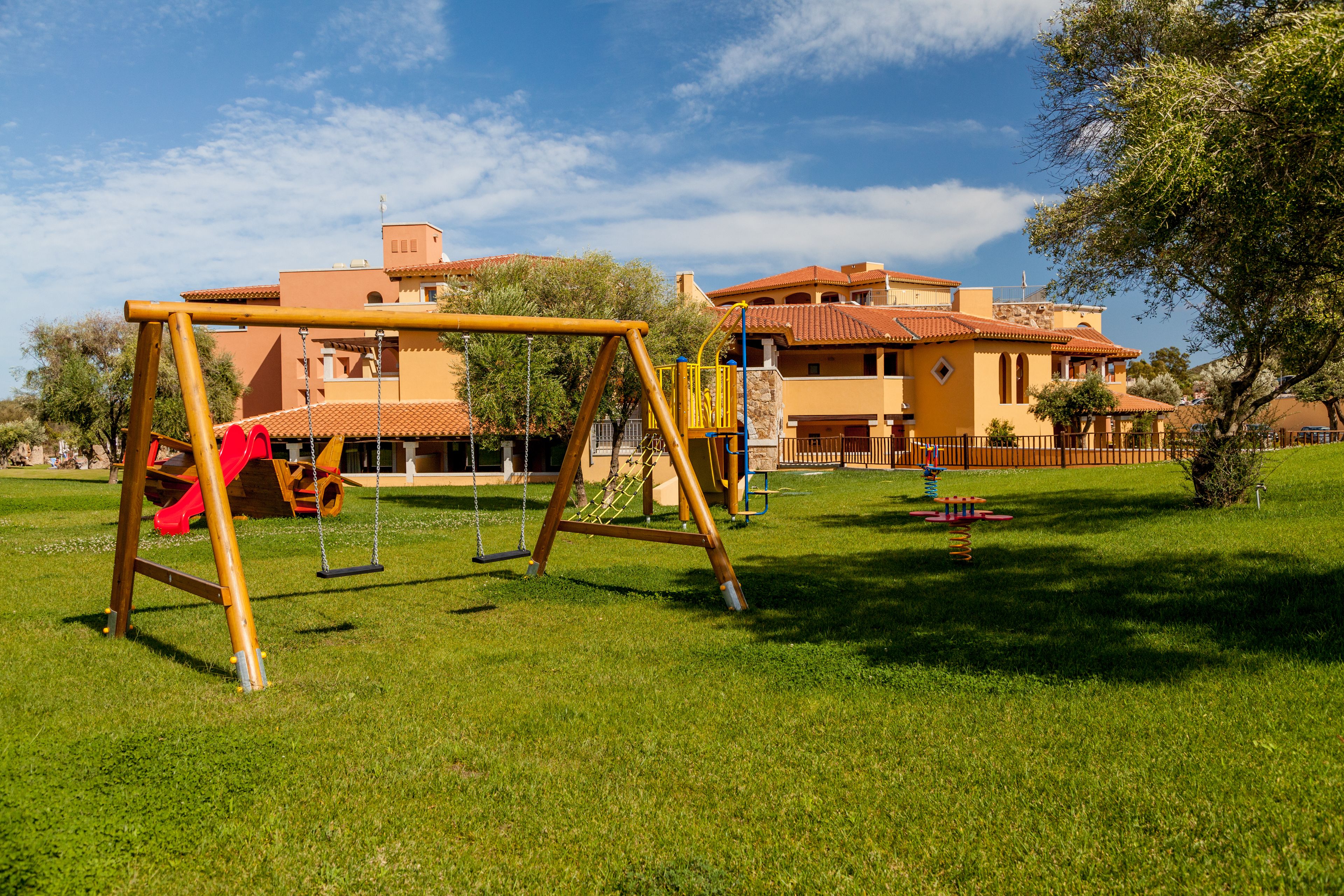Un parque infantil con columpios y un tobogán, rodeado de césped cuidado y un edificio con techo de tejas.