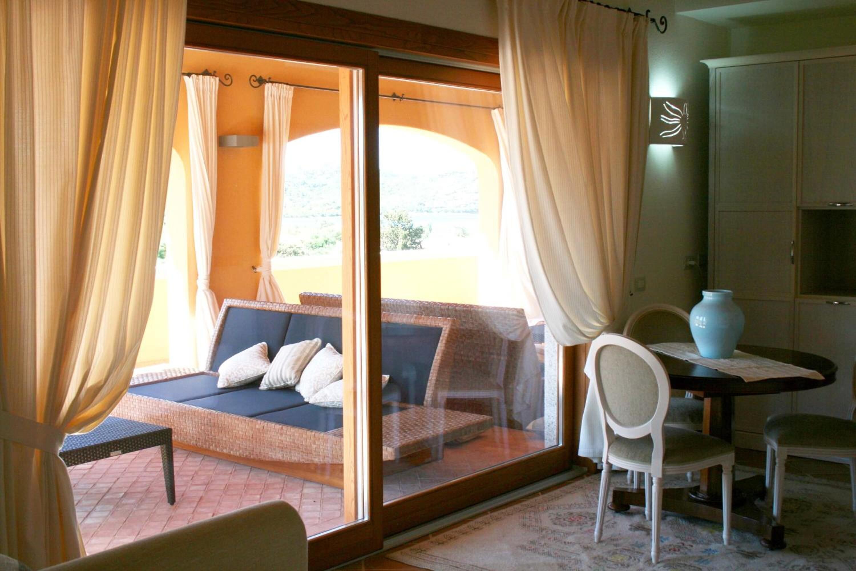 A room with a sofa bed, a dining table with chairs, a vase, and a view through a glass door.