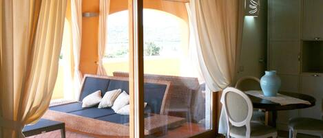 A room with a sofa bed, a dining table with chairs, a vase, and a view through a glass door.