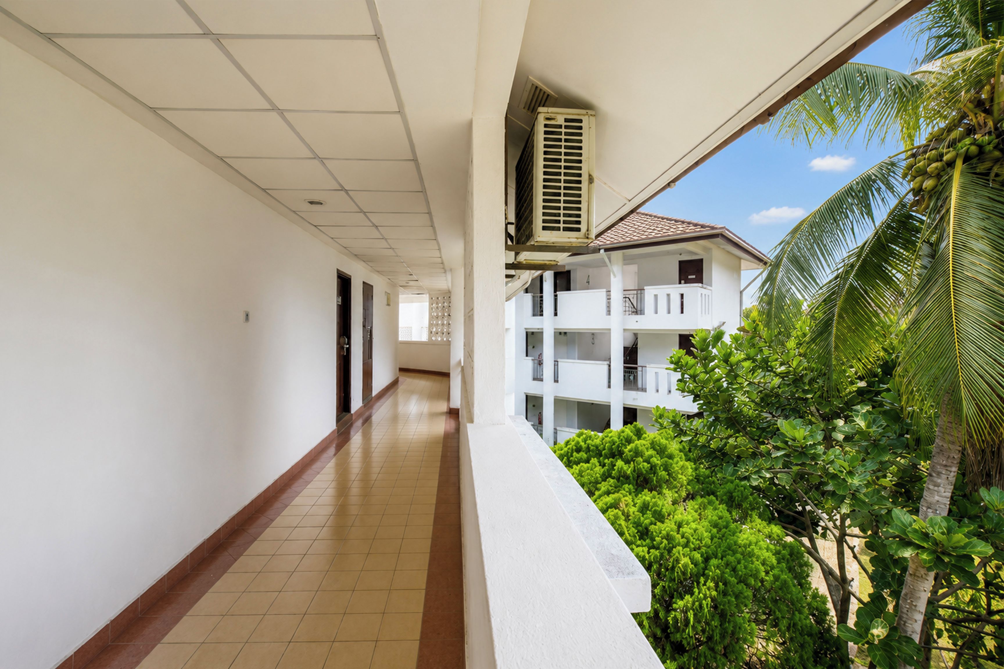 A corridor with a tiled floor, white walls, and a view of a building with balconies and greenery.
