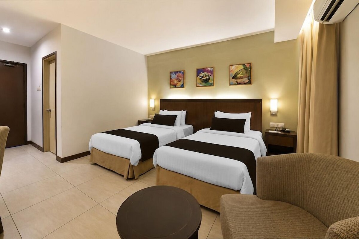 A hotel room with two beds, a small round table, and a sofa. There are wall-mounted lamps and artwork above the beds.