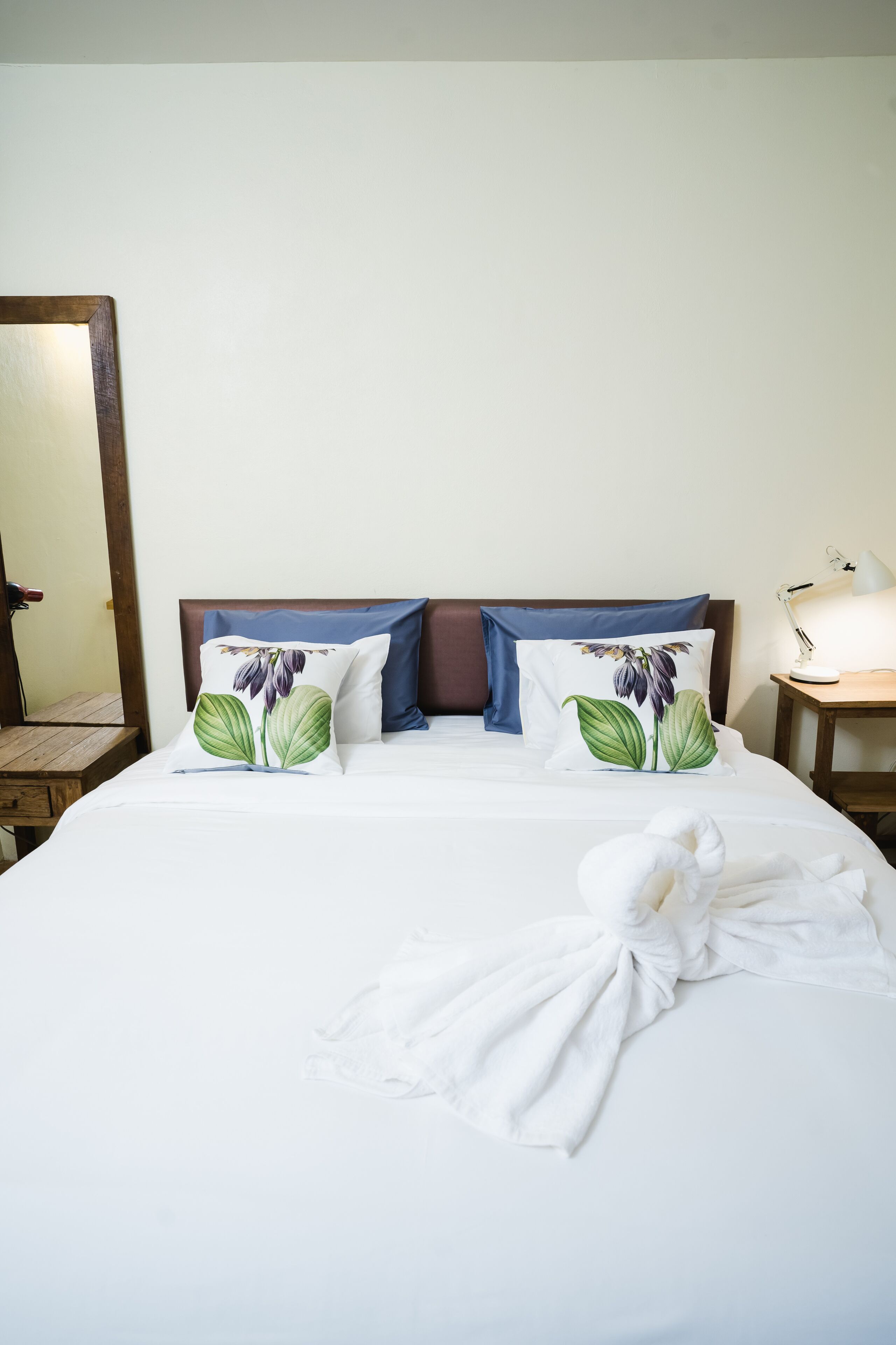 Una cama bien hecha con almohadillas florales, un cabecero de madera, una lámpara de noche y un espejo en la pared.