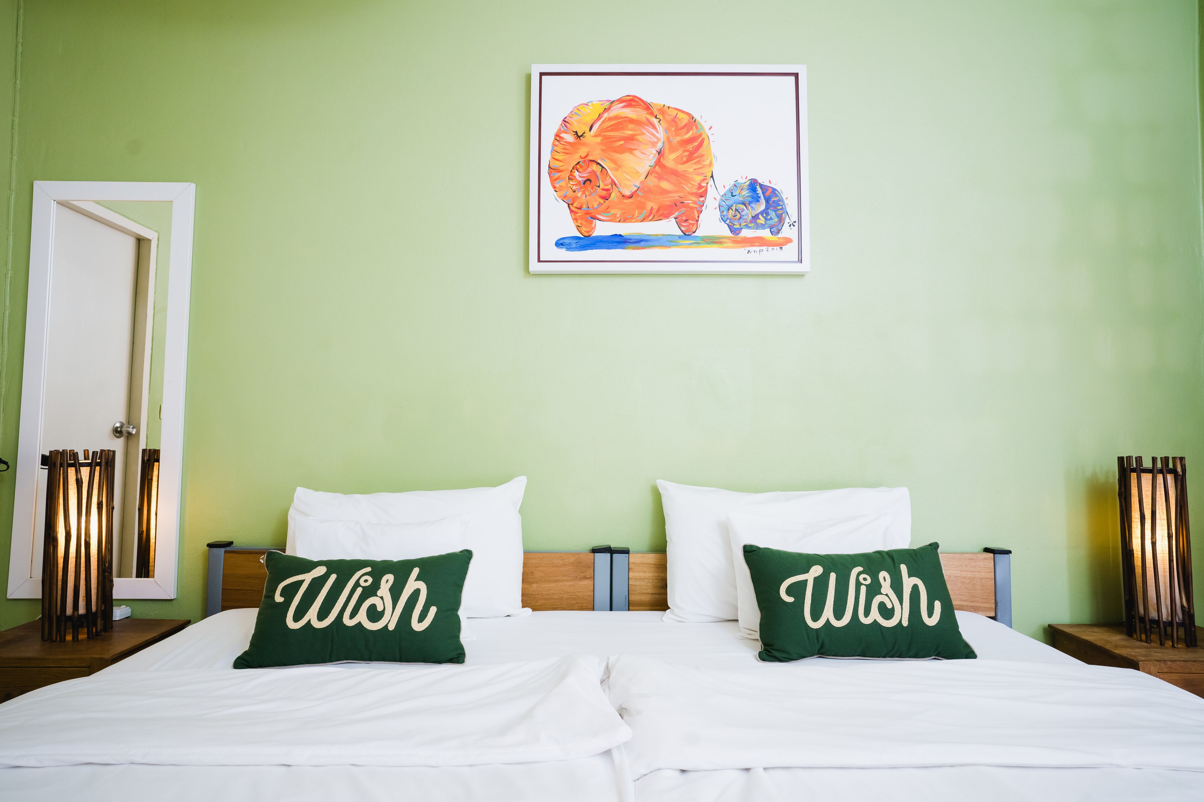 Una habitación de hotel con una cama, dos almohadas con la palabra "Wish" y una obra de arte enmarcada sobre la cama, y una puerta a la izquierda.