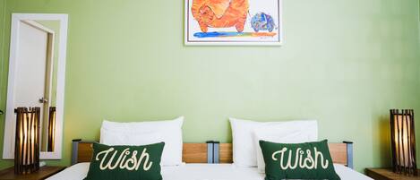 Una habitación de hotel con una cama, dos almohadas con la palabra "Wish" y una obra de arte enmarcada sobre la cama, y una puerta a la izquierda.