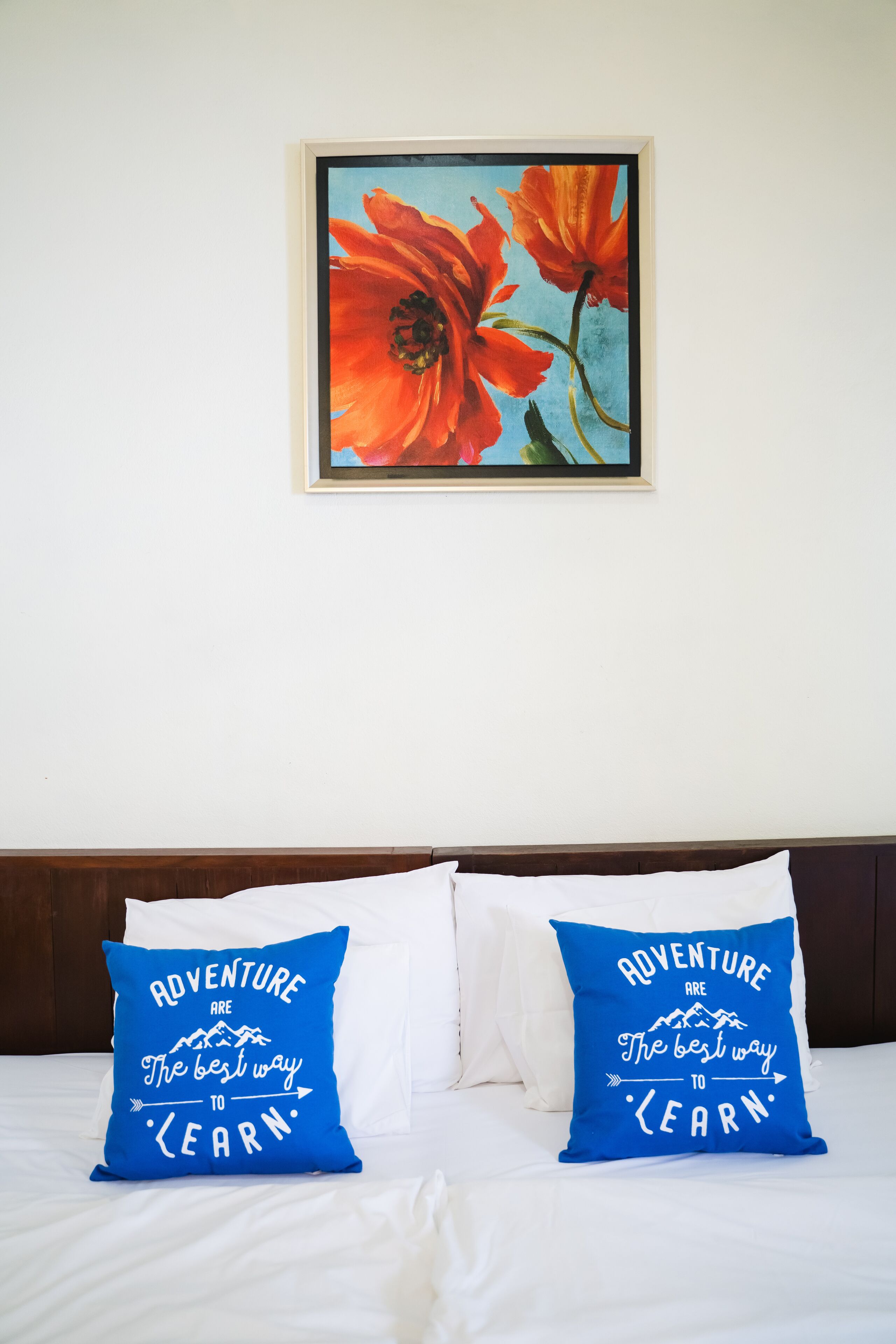 A bed with white bedding and two blue pillows with motivational text, a framed painting of red flowers above the bed, and a wooden headboard.