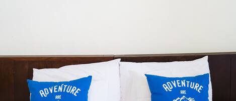 Una cama con ropa blanca y dos almohadas azules con texto motivacional, un cuadro con flores rojas sobre la cama, y un cabecero de madera.